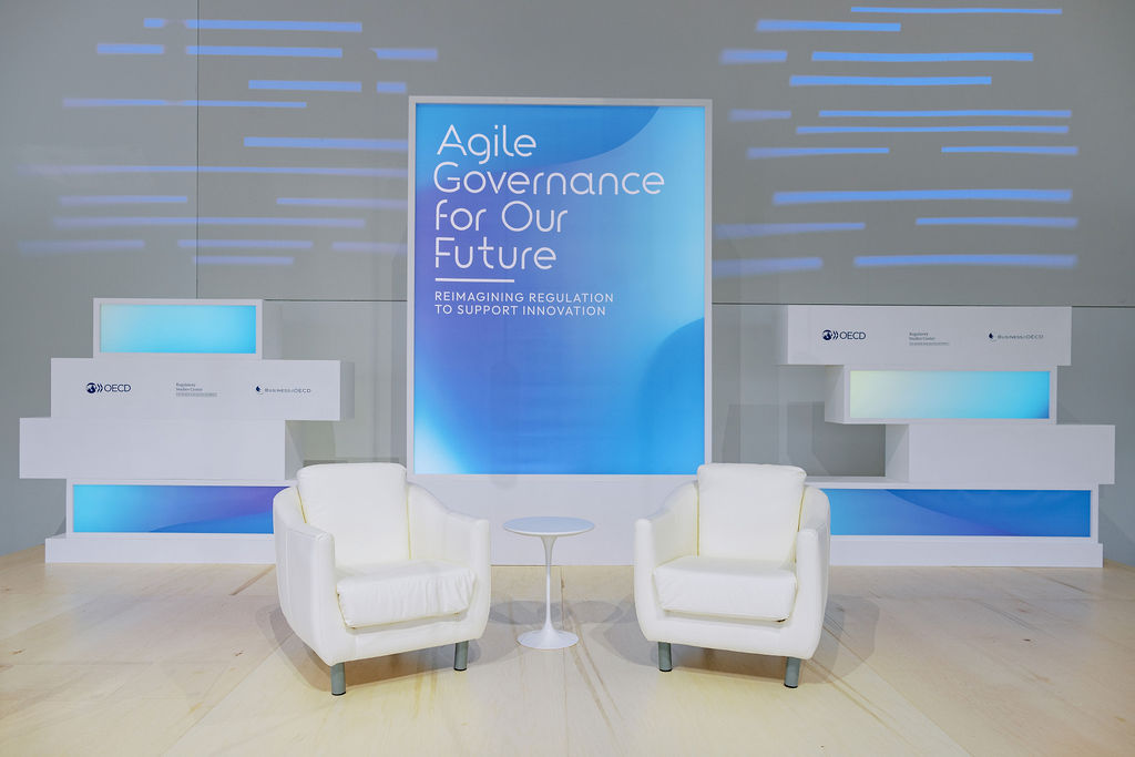 Two chairs on a stage in front of sign reading "Agile Governance for our Future: Reimagining Regulation to Support Innovation"