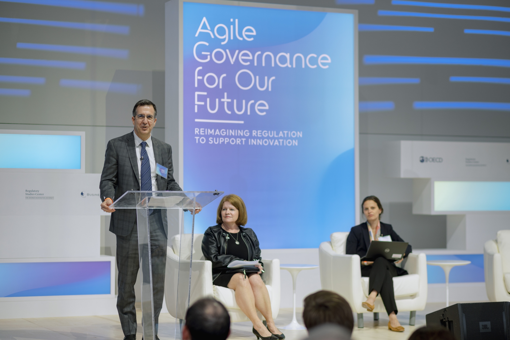 Dom Mancini speaking at a lectern with two others seated on stage