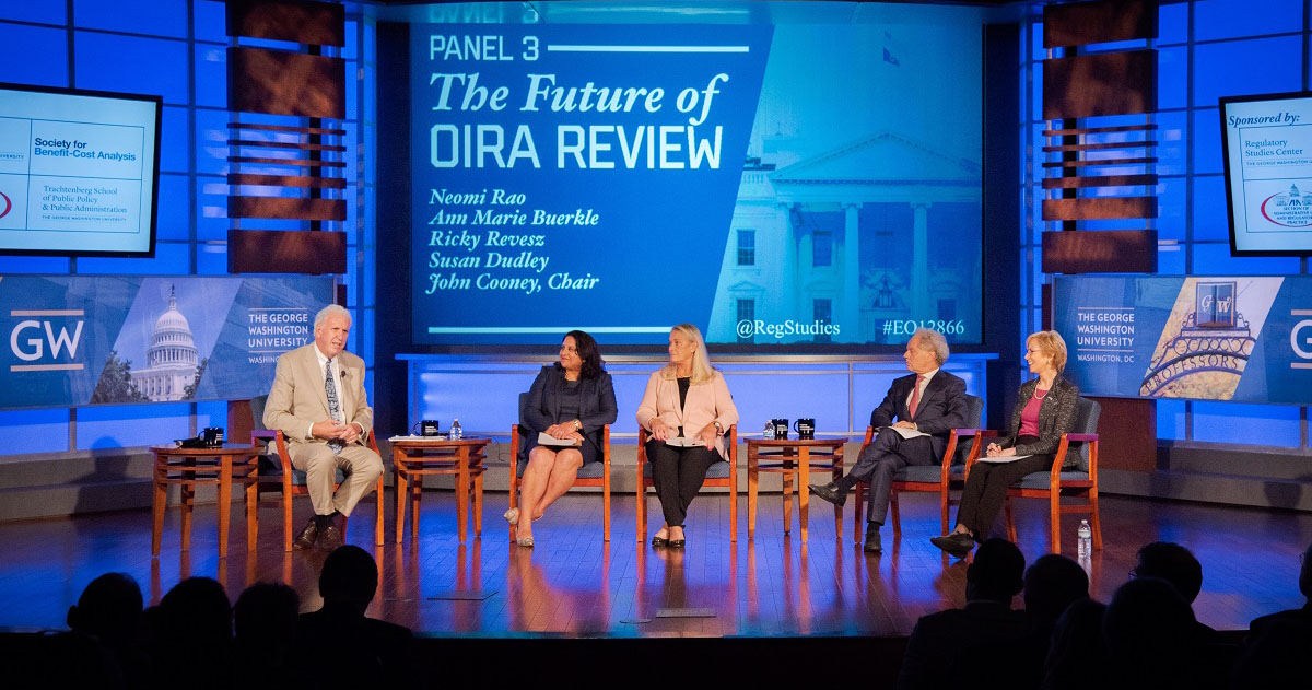 Panelists sitting on stage at a Reg Studies event with a screen: The Future of OIRA Review
