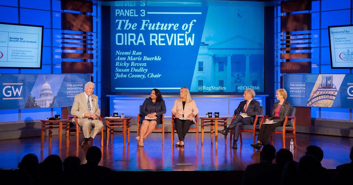 Panelists sitting on stage at a Reg Studies event with a screen: The Future of OIRA Review
